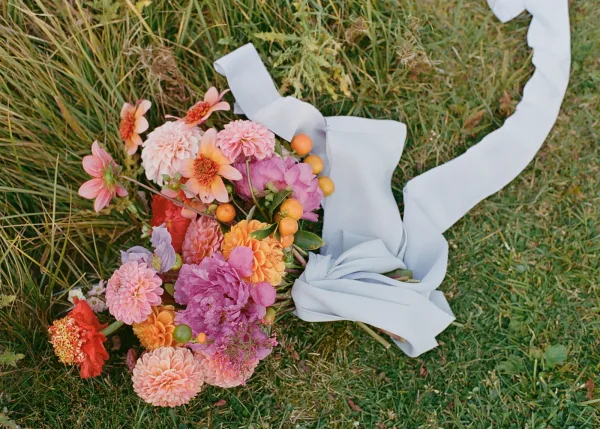 Bridal bouquet with colorful wedding bouquet blooms—dahlias, peonies, poppy, berry stems and greenery—wrapped in a white ribbon on meadow grass