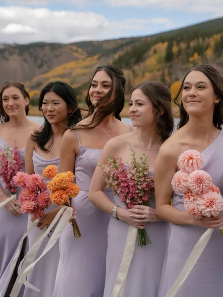 Bridesmaid portraits of women holding bouquets in lavender bridesmaid dresses with ribbon streamers by a mountain lake under cloudy autumn skies