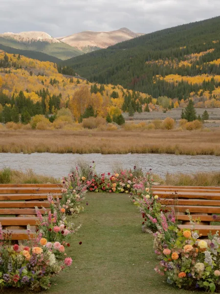 Ceremony setup with floral aisle arrangements lining a grass runner, wooden benches facing a lakeside meadow with mountains and autumn trees under cloudy sky
