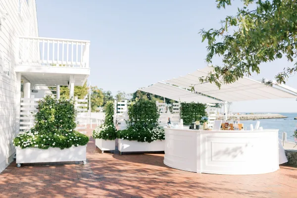 Wedding bar setup for an outdoor cocktail hour, featuring blue hydrangea, on a brick patio by the waterfront with trees nearby.