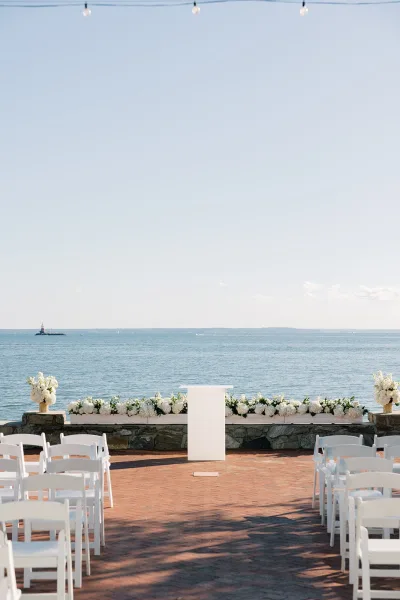 Ceremony setup for a waterfront ceremony with white florals framing a simple altar on a brick terrace, backed by open water and sky.