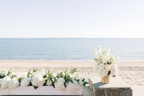 Ceremony flowers styled as beach ceremony decor with white hydrangeas by the ocean and sandy beach beside a low stone wall under clear sky