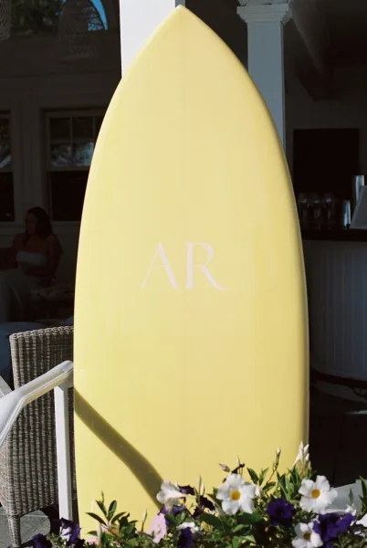 Wedding signage styled as a surfboard wedding sign, featuring bold monogram lettering by a bar counter with white paneling and lanterns.