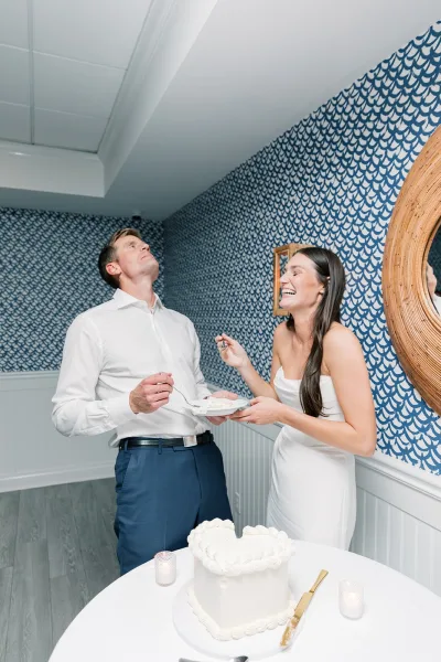 Wedding cake cutting as a couple feed each other cake, with a gold cake knife beside a square white cake against blue patterned wallpaper.