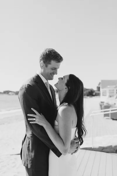 Couple portrait of a beach wedding couple embracing on a boardwalk, her engagement ring catching light as houses dot the shoreline behind.