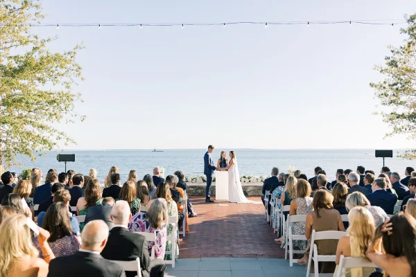 Ceremony moment during waterfront wedding vows as the bride and groom stand at the altar, her long veil catching light, framed by ocean and clear sky.