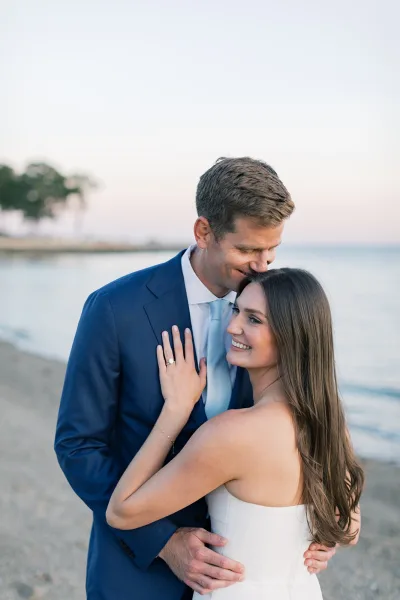 Couple portrait as a bride and groom embrace on a sandy beach, groom in a blue suit, shoreline water and distant trees behind them.
