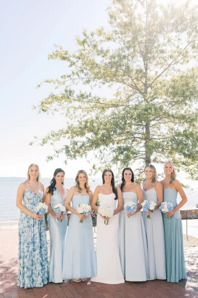 Bride and bridesmaids in blue stand on a brick patio by the waterfront, holding bouquets beside a leafy tree.