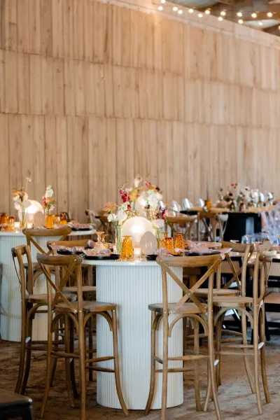 Reception tablescape with amber glass goblets, pink napkins, and floral centerpieces on a round bar-height table under string lights by a wood wall