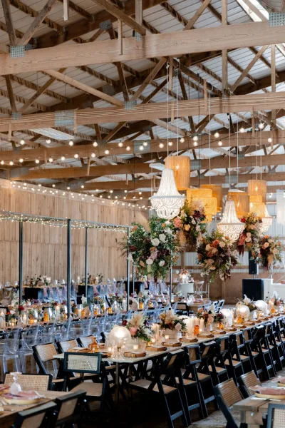 Reception tablescape with a long banquet table wedding setup, candlelit florals, amber goblets, and chandeliers under barn beams with string lights