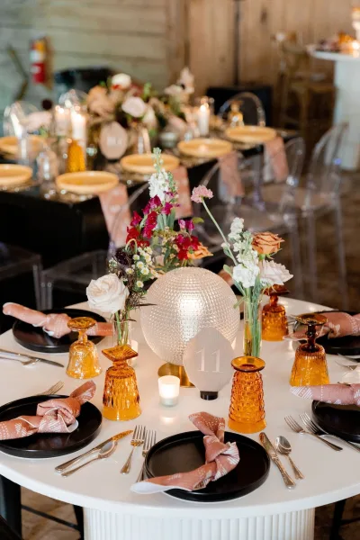 Reception tablescape with round wedding table setting, black plates and amber goblets, floral centerpieces and candles on white linens by wood walls