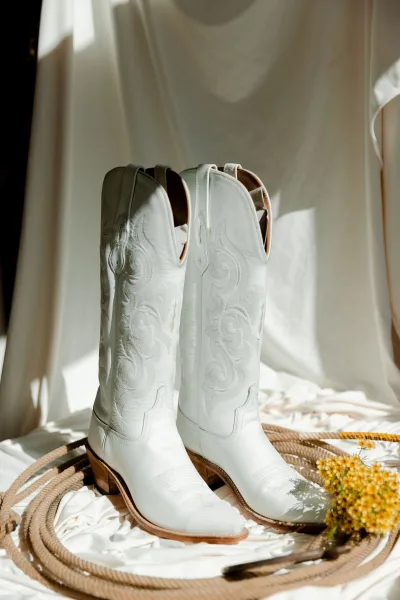 Wedding boots with white cowboy boots in embroidered leather beside coiled rope and yellow flowers on a sunlit white fabric backdrop
