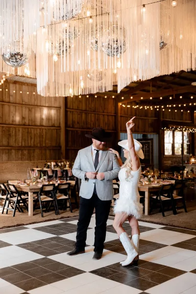 Wedding first dance as bride and groom in cowboy hats sway on a checkered dance floor under disco balls and string lights in a barn