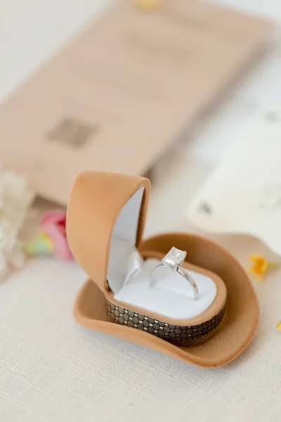 Engagement ring with a diamond solitaire ring in an open ring box on linen fabric, beside blurred invitation stationery and petals