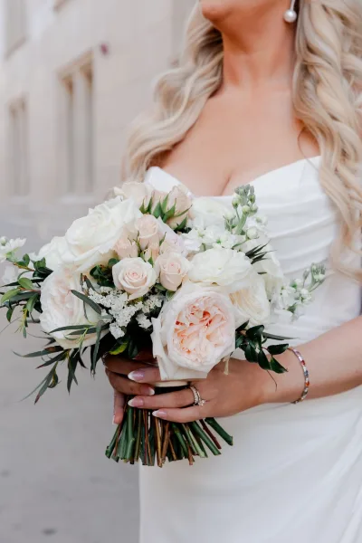 Bridal bouquet of white and blush roses with greenery, held by a bride in a strapless gown with pearl earrings against a neutral wall