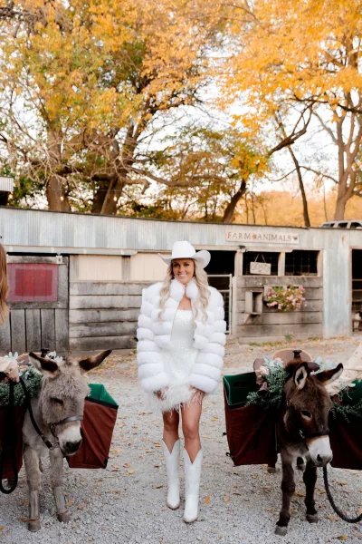 Bridal portrait of a cowgirl bride look in a mini wedding dress and white cowboy hat, standing by donkeys near a rustic barn in fall