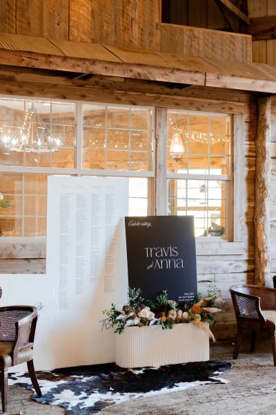 Wedding seating chart sign displayed on a plinth with greenery and pampas grass accents, set against a rustic wood wall with windows and string lights