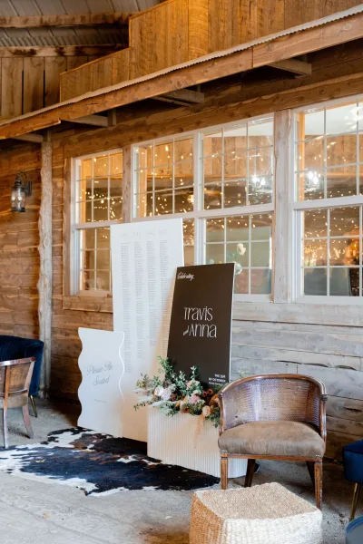 Wedding seating chart display on a board beside a welcome sign with floral arrangement, set against a rustic barn wall with windows and string lights.