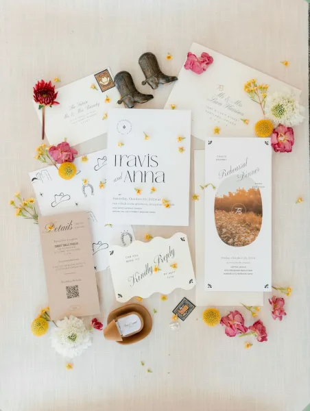 Invitation suite wedding invitation flat lay with envelopes, RSVP and details cards, postage stamps, flowers, ring box, and cowboy hat on linen backdrop