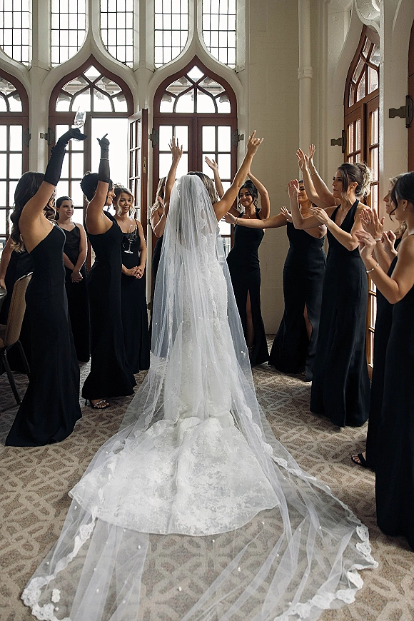 Bride with bridesmaids cheering during a bridesmaids reveal moment, lace train and veil flowing as they raise champagne in a hallway