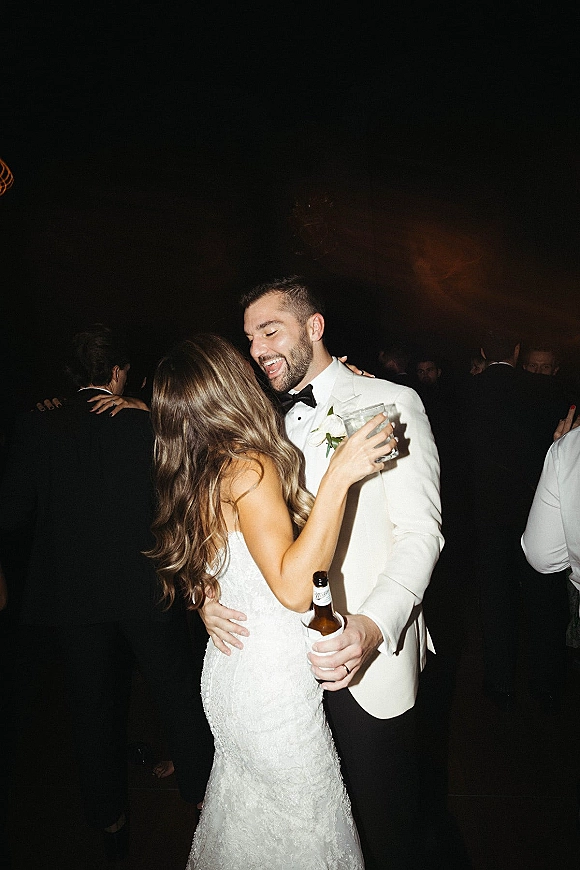 Wedding reception dancing as bride in lace dress holds a cocktail and groom in white tuxedo with beer dances close in dim crowd