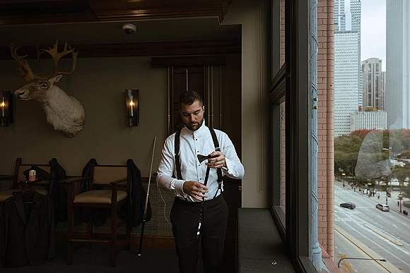 Groom getting ready, adjusting bow tie with black suspenders over a white dress shirt in a hotel room by a large window and skyline view