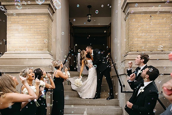 Wedding kiss as newlyweds dip on stone steps while bridal party blows bubbles, bride in strapless lace gown and cathedral veil