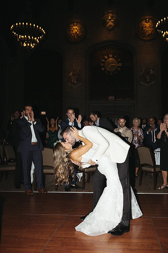 Wedding first dance dip kiss as bride in lace wedding dress and groom in white dinner jacket sway under chandelier lighting in a ballroom
