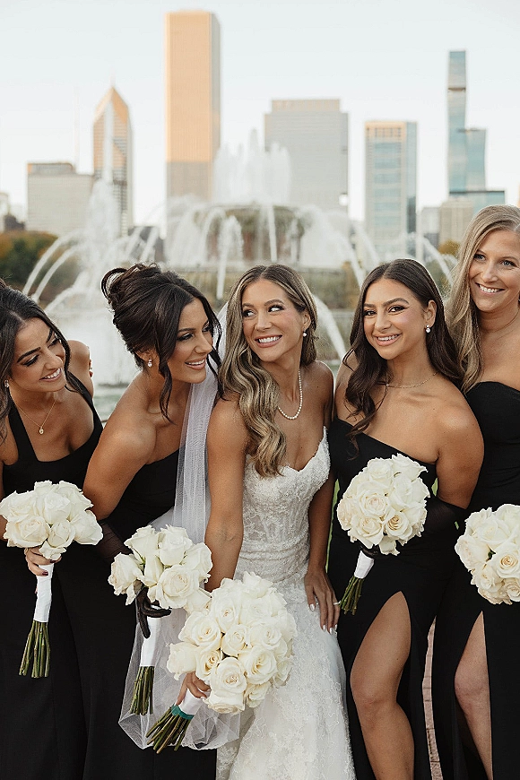 Bridesmaid group photo with bride and bridesmaids holding white rose bouquets, laughing by a fountain with a city skyline backdrop