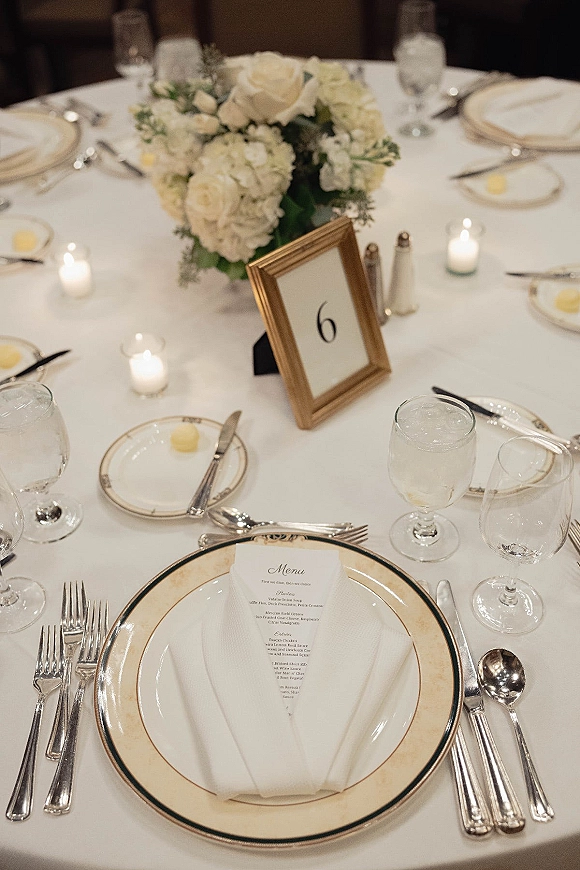 Reception tablescape wedding table setting with white hydrangea and rose centerpiece, votive candles, gold table number, and formal place settings