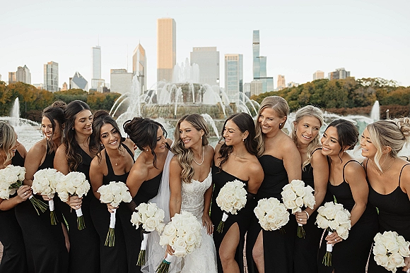 Bridesmaid group photo with bride with bridesmaids in black dresses holding white rose bouquets by a fountain, city skyline behind them