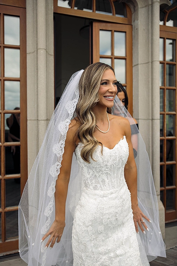 Bridal portrait of a bride in a strapless lace wedding dress, smiling by wooden French doors, lace veil and pearl necklace visible