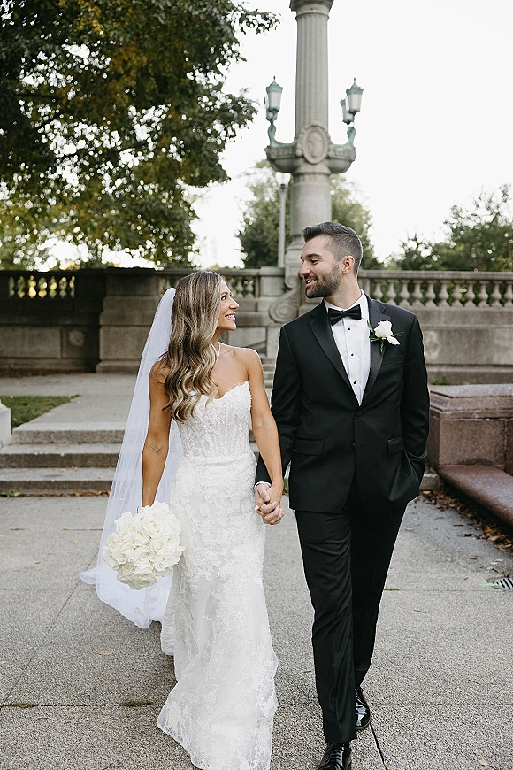Couple portrait of bride and groom walking hand in hand, she holds a white bouquet and veil trails beside stone steps and trees