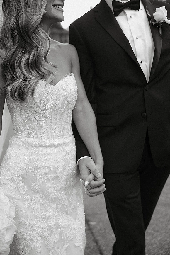 Bride and groom holding hands in a black and white portrait, her strapless lace dress and bouquet beside his tuxedo and boutonniere outdoors
