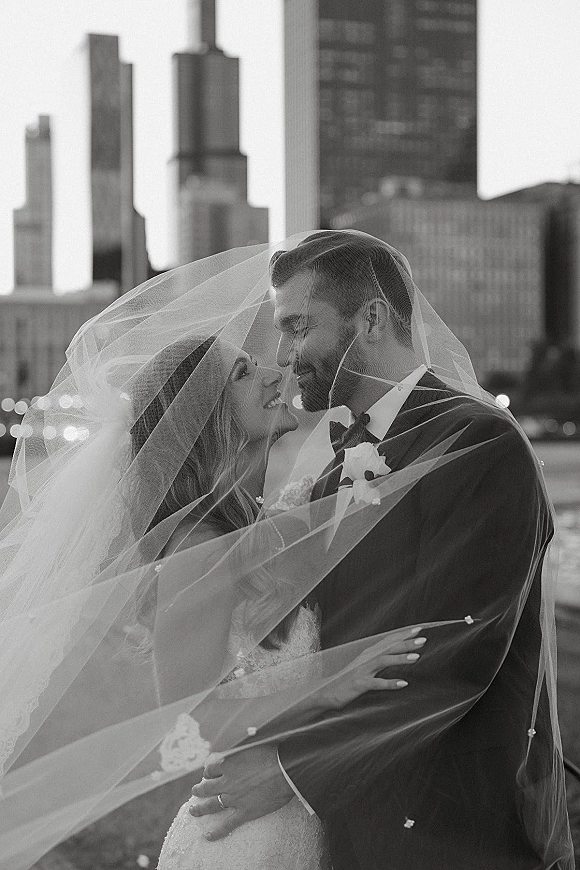 Wedding couple portrait of bride and groom under veil, foreheads touching, tuxedo and lace dress with skyline and waterfront behind