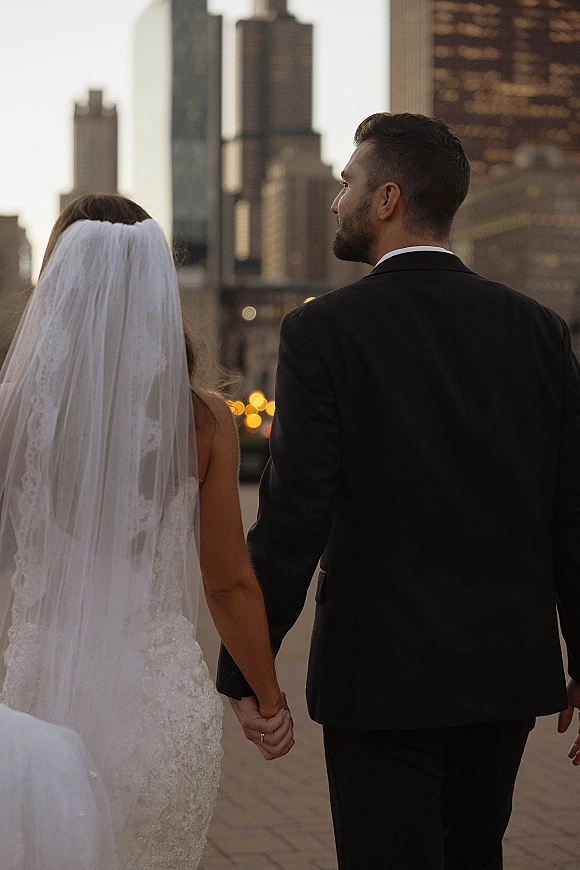Couple portrait of bride and groom holding hands, walking away in a lace dress and veil, with evening city skyline lights behind