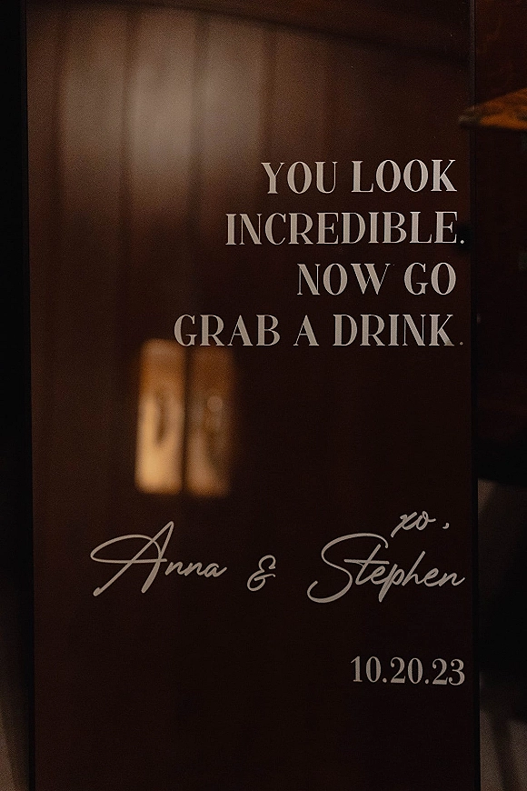 Wedding welcome sign on clear acrylic with white calligraphy lettering, displayed against a dark doorway and interior wall backdrop