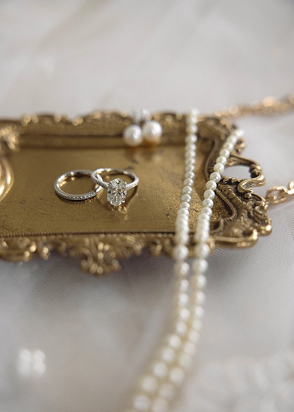Wedding rings with a diamond engagement ring stacked on a band, styled on a vintage gold tray with pearls on white fabric