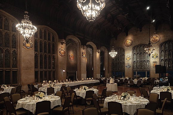 Wedding reception decor with round tables in white linens, floral centerpieces and candles beneath chandeliers in a historic stone hall