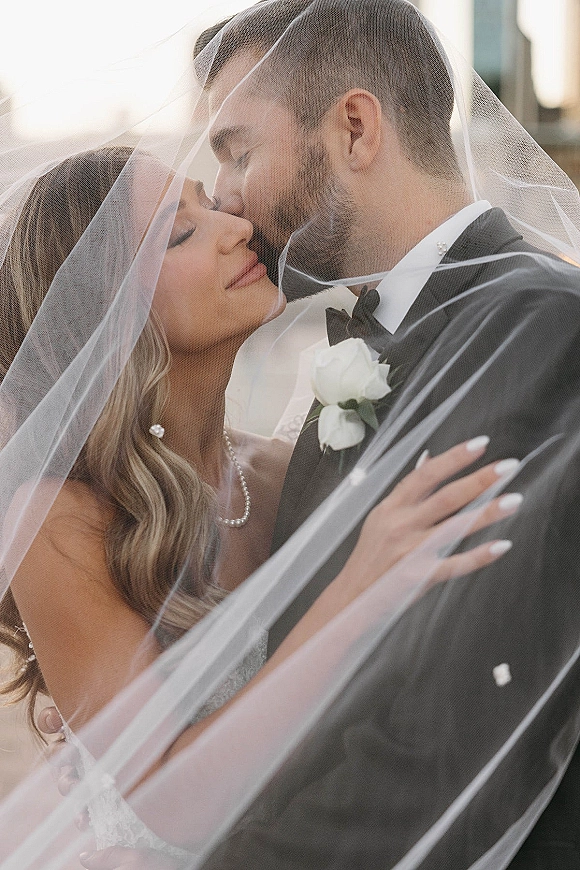 Wedding kiss as groom kisses bride’s forehead under a sheer veil, her hand on his shoulder, in an outdoor setting near a building