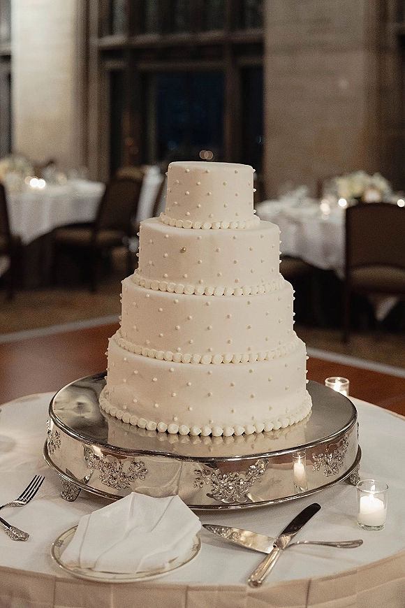 Wedding cake with white fondant, four tiers, pearl piping and dot-texture icing on a silver stand with votive candles in reception room