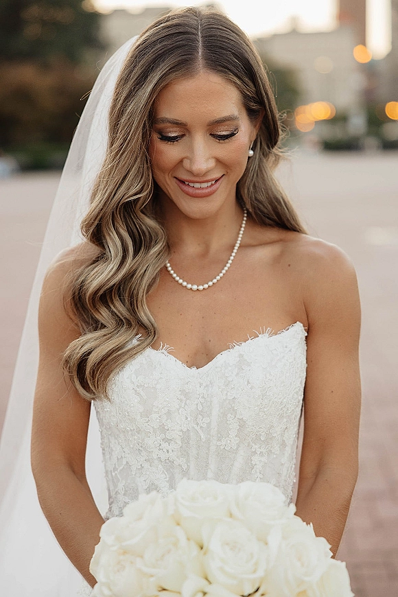 Bridal portrait of a bride looking down, holding a white rose bouquet in a strapless lace dress with veil on an outdoor walkway with bokeh lights