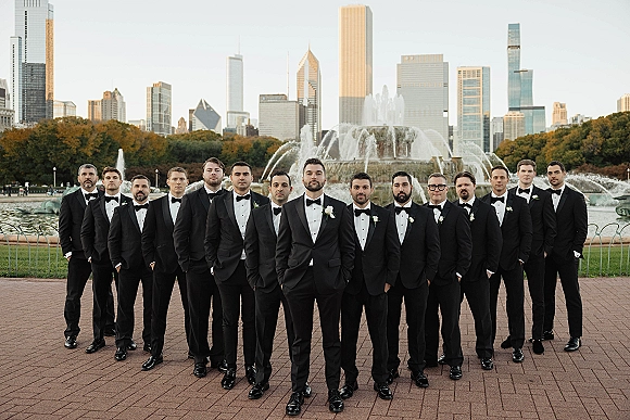 Groomsmen portrait of a lineup in black tuxedos and bow ties on a brick walkway by a fountain, with city skyline behind them
