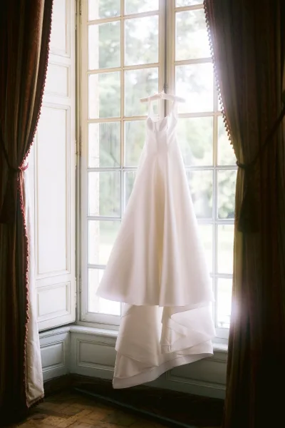 Wedding dress, a simple wedding dress with an ivory A-line V-neck and thick straps, hangs on a hanger before a large window framing garden trees.