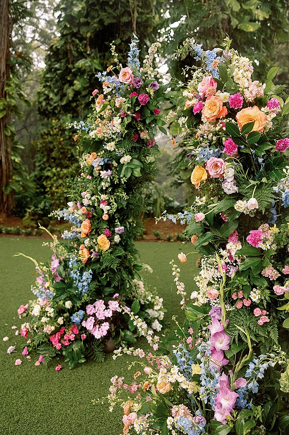 Floral wedding arch with roses, hydrangea, and delphinium in a garden ceremony arch, lush greenery and hanging vines on a lawn with trees
