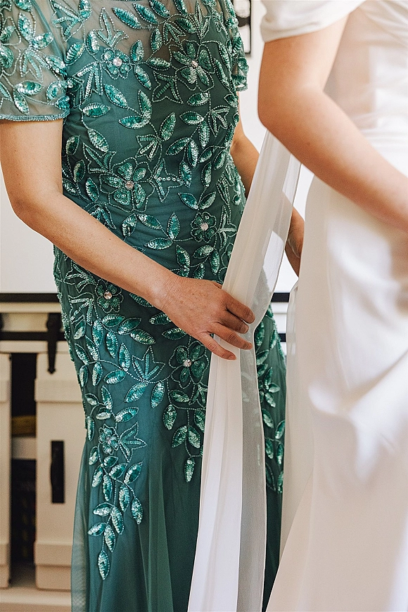 Bridesmaid dress in emerald green with beaded embroidery and sequins, chiffon sheer overlay detail hanging in an indoor room by cabinetry