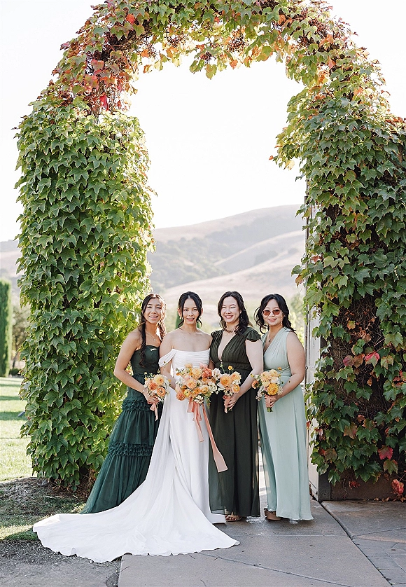 Bridesmaid group portrait with bride and bridesmaids in mixed green dresses holding peach and yellow bouquets, under an ivy archway with mountains behind