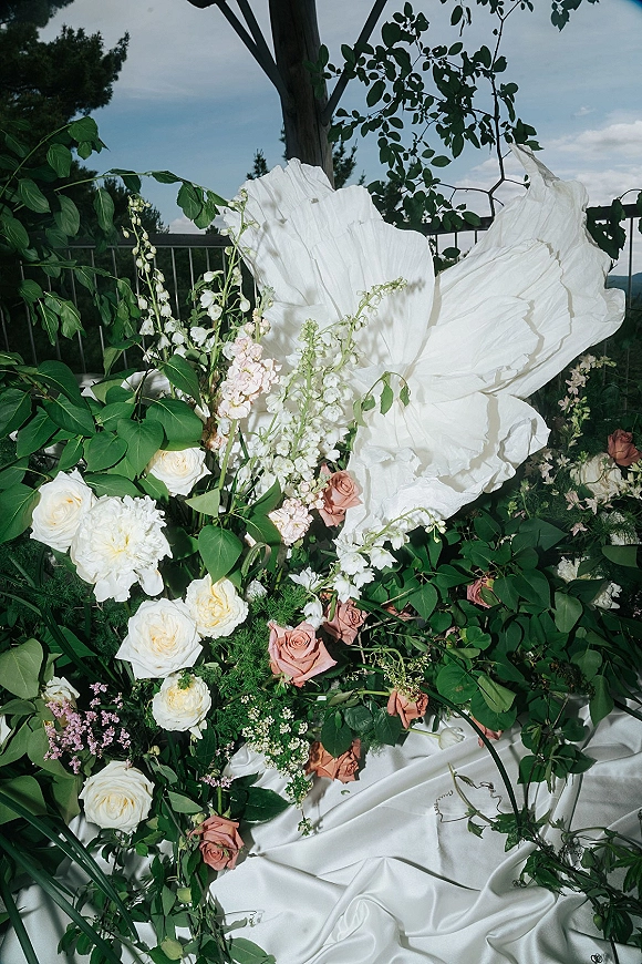 Wedding floral arrangement with white and blush blooms, pink roses, and greenery, set beside draped fabric and a large paper flower on an outdoor terrace
