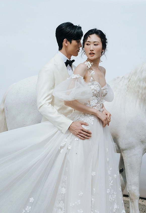 Couple portrait of groom hugging bride in an off-the-shoulder puff-sleeve tulle gown, beside a white horse against open sky backdrop
