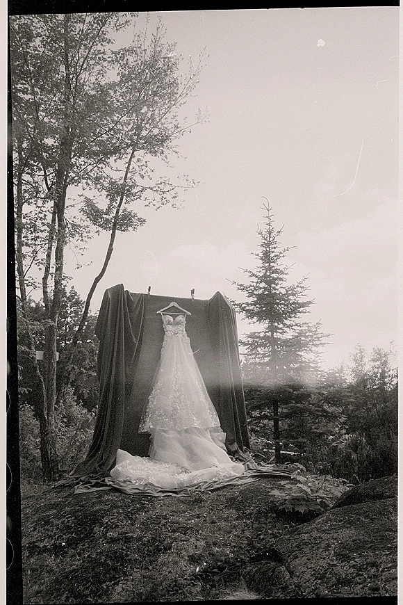 Wedding dress with lace overlay and sweetheart neckline hanging on a hanger, framed by draped cloth with forest trees and rocks behind